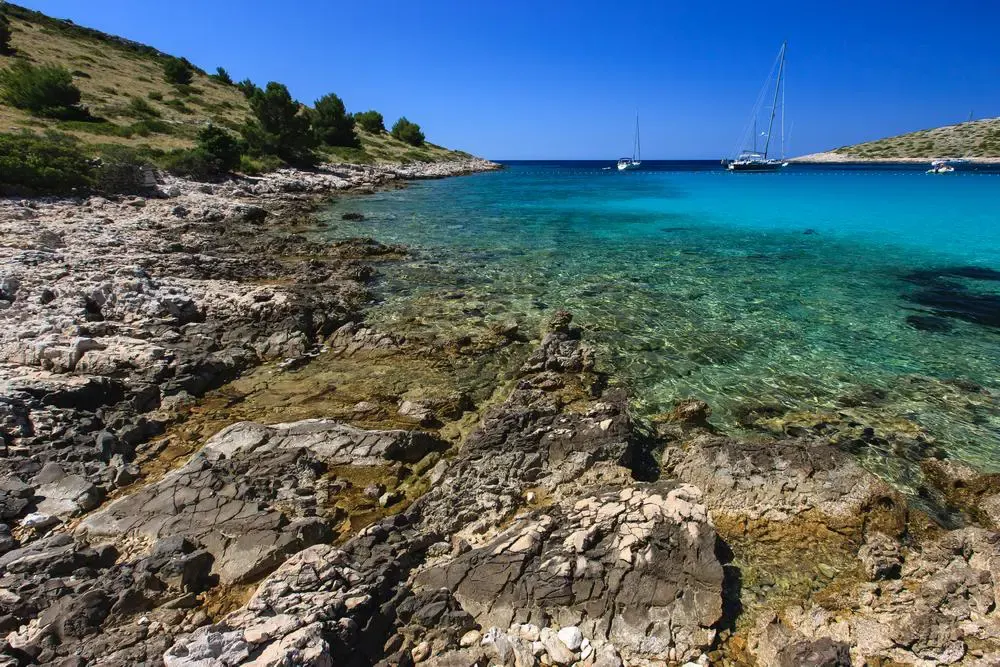 Sailing yachts anchored in a clear turquoise cove on a rocky Kornati shore