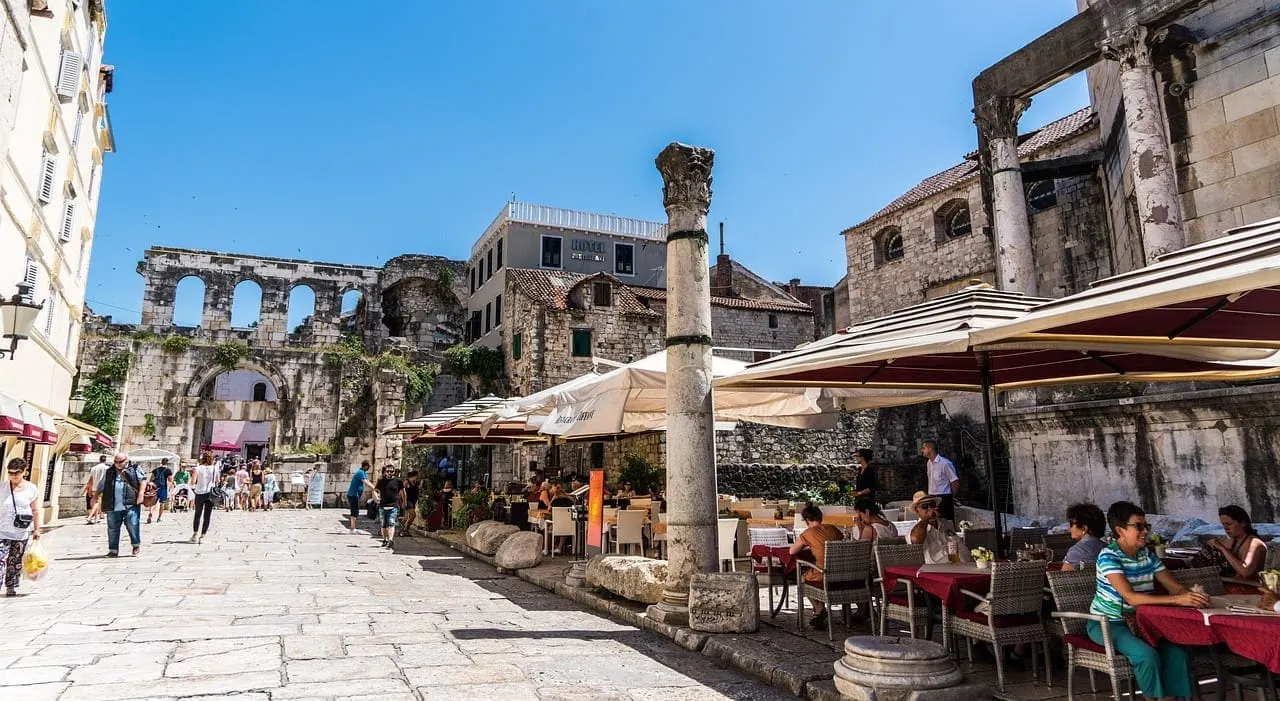 Historic stone ruins and Roman columns frame a Mediterranean waterfront square with outdoor café seating and visitors