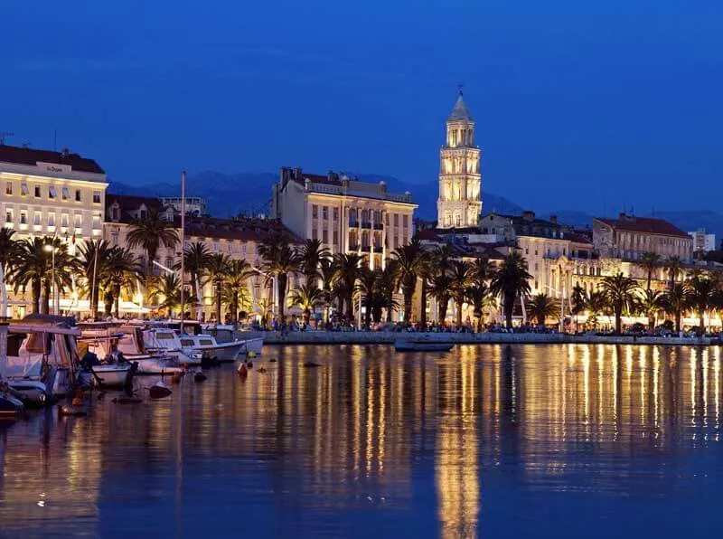Fishing boats moored at dusk in a Mediterranean harbour with illuminated waterfront buildings and a bell tower reflected in calm water