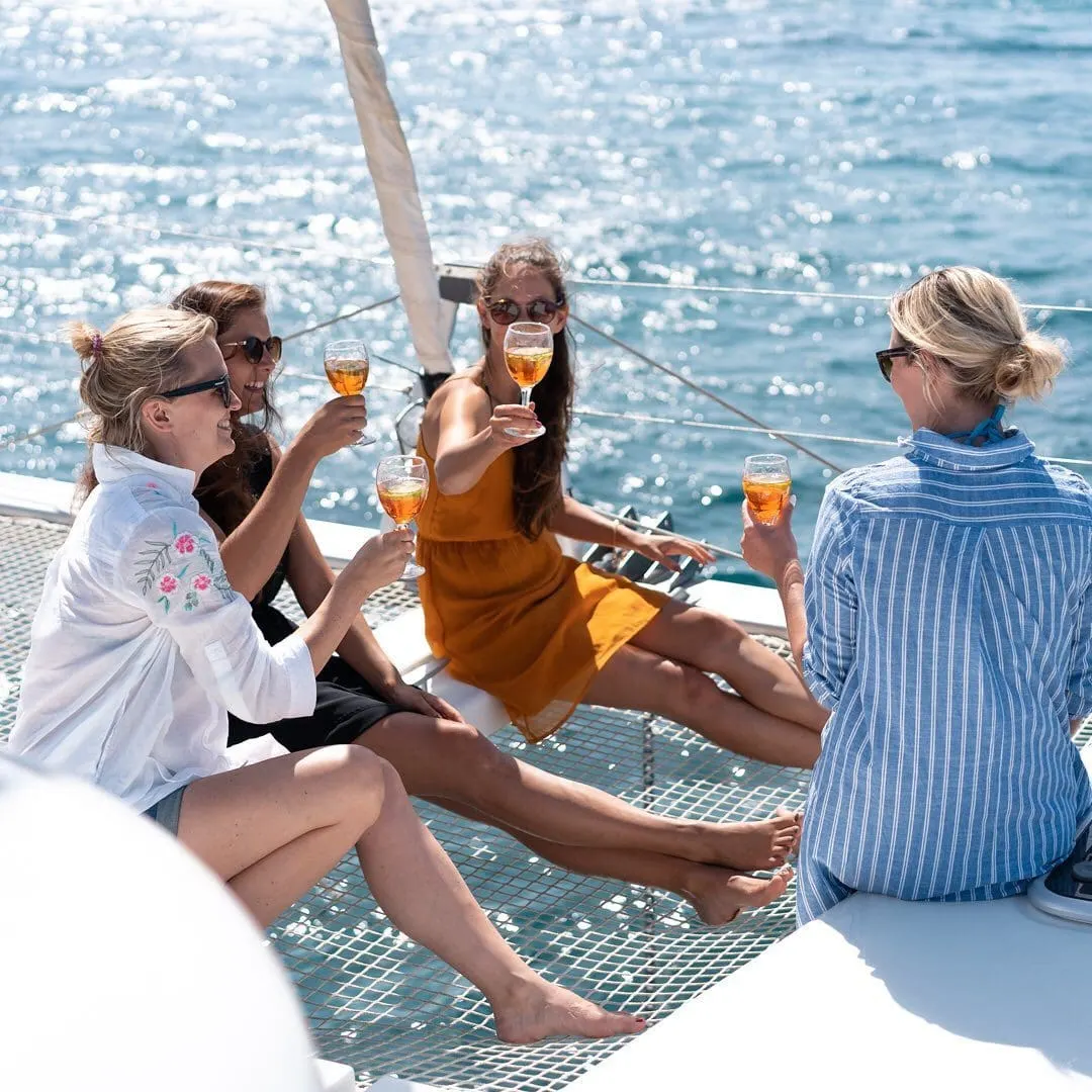 Four friends in light summer clothes and sunglasses raising drinks on a catamaran trampoline