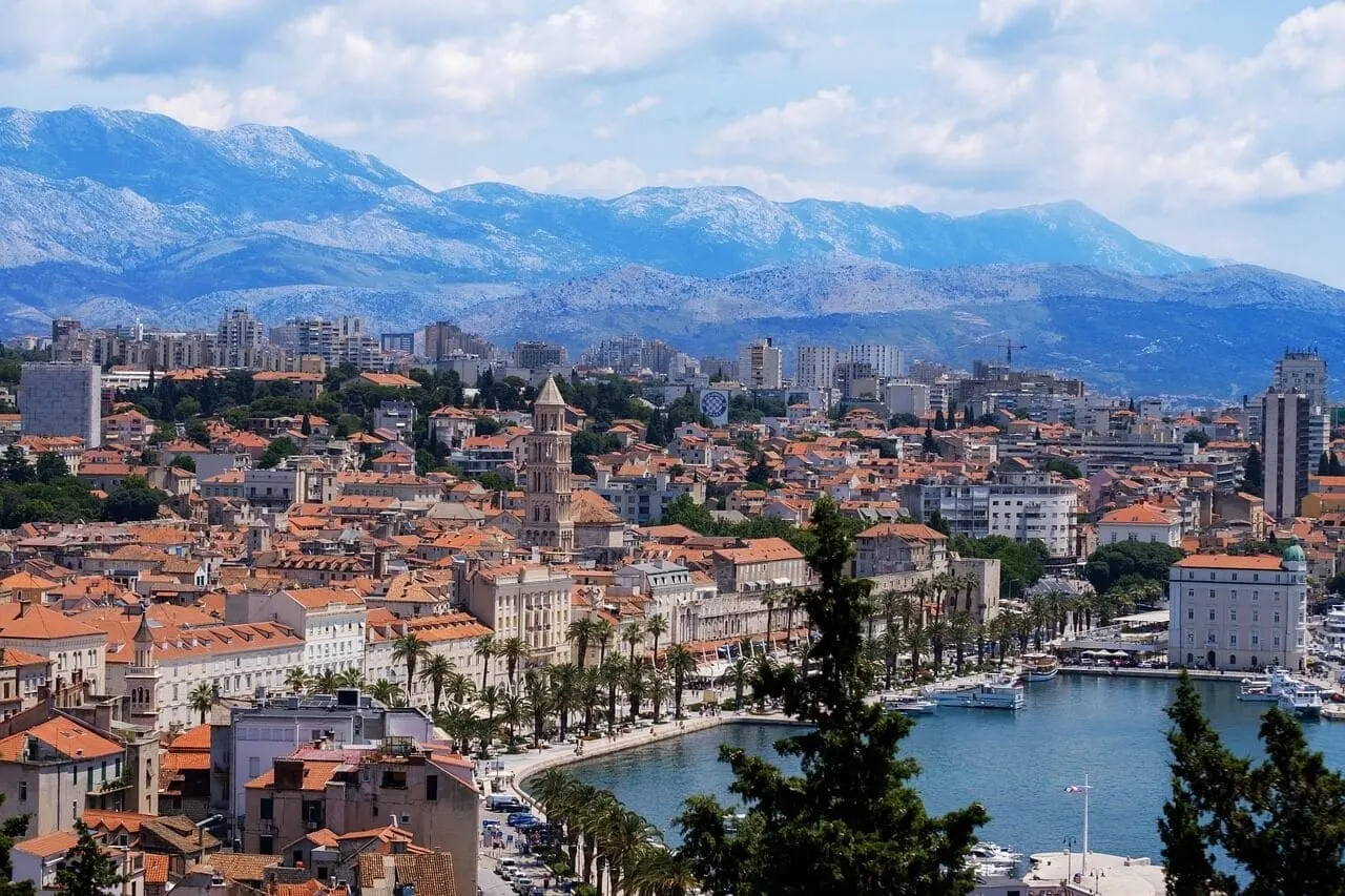 Coastal city with terracotta roofs, waterfront promenade with palm trees, and mountains rising behind, moored yachts visible in harbor